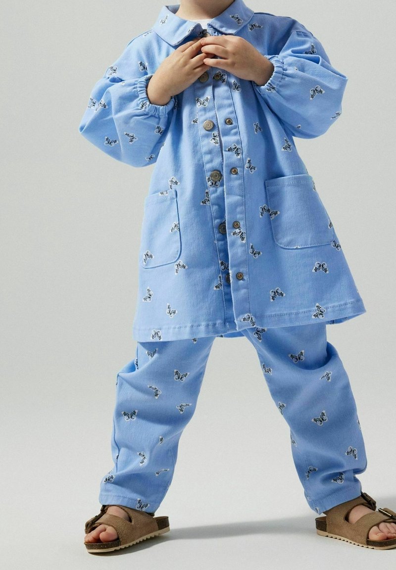 Enfant portant une tenue bleue boutonnée avec des motifs de papillons et des sandales marron, debout jambes écartées et mains près du col.