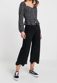 Blouse à carreaux noir et blanc avec des manches longues et une fermeture à boutons, associée à un jean noir à jambes larges avec des bords effilochés et des baskets noires.