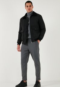 Črna bomber jakna iz volnenega mešanice z drugim slojem ovratnika, dvema sprednjima žepoma in rebrastimi manšetami, v kombinaciji s sivo krojenimi hlačami in črnimi športnimi copati.