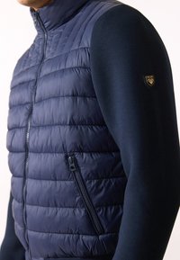 Marinblå pufferväst med quiltat mönster, har en främre dragkedjeficka och åtsittande ärmar i solid marinblått tyg. Inkluderar en logodetalj.