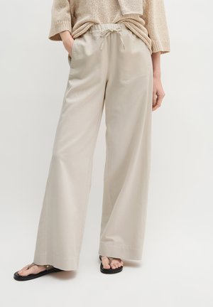 Pantalon beige à jambes larges avec taille à cordon, porté avec un pull beige en maille ample et des sandales plates noires sur fond blanc.