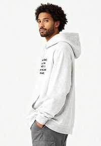 Felpa con cappuccio grigio chiaro in tessuto felpato con tasca frontale. Presenta testo nero sul lato sinistro del petto. Texture morbida e vestibilità rilassata.