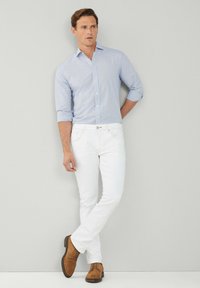 Camicia button-up a scacchi blu chiaro con maniche arrotolate, jeans slim-fit bianchi e scarpe in camoscio marroni, in piedi davanti a uno sfondo grigio.