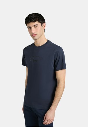 Giovane uomo con una maglietta a maniche corte blu navy scuro con un logo in rilievo sottile, in piedi contro uno sfondo chiaro uniforme.