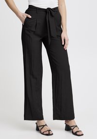 Pantalons larges noirs avec une finition texturée, dotés d'une taille nouée, de poches avant et d'une coupe décontractée, associés à des sandales à talons carrés noirs.