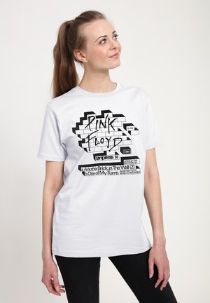 Jeune femme avec queue de cheval brune portant un T-shirt blanc Pink Floyd avec un graphisme de mur en briques et titres de chansons, debout devant un fond uni.