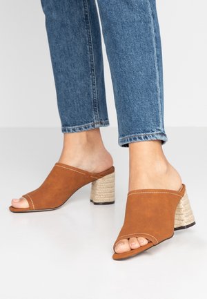 Mules ouverts en daim marron avec une tige incurvée et un talon bloc texturé de style tissé. Surface lisse et détails de couture contrastants.