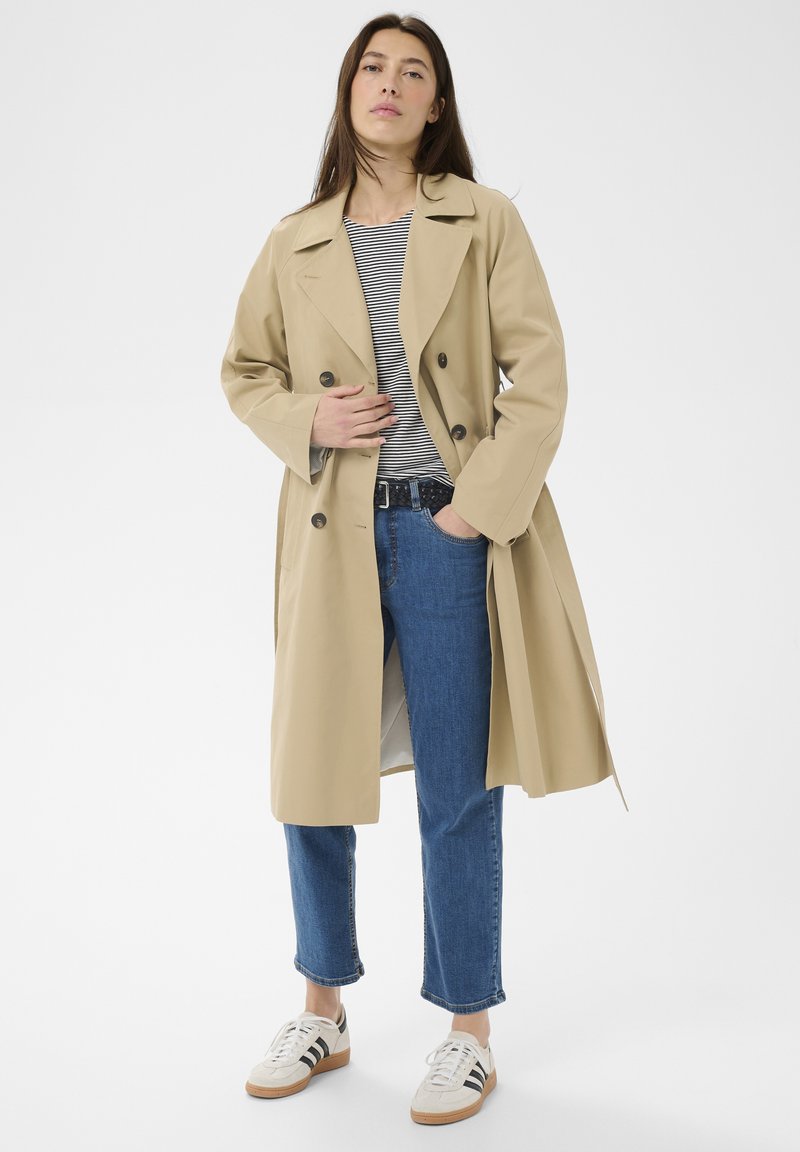 Femme portant un trench-coat beige, une chemise à rayures, un jean bleu et des baskets blanches à rayures noires, debout avec une main dans la poche.