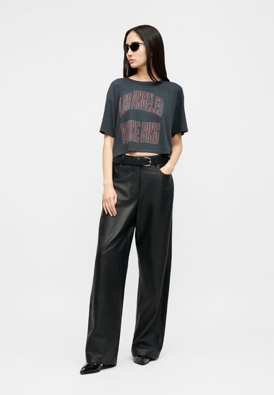 Nő fekete napszemüvegben, sötétszürke, rövidített "Los Angeles Aime Bing" feliratú pólóban, bőszárú fekete bőrnadrágban és fekete hegyes orrú cipőben.