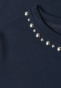 Dark blue fabric with round silver studs arranged in a symmetrical pattern along the neckline edge of a garment.