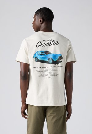 Férfi, aki krémszínű pólót visel kék vintage autós grafikával és a"Denim Gremlin" felirattal a háton, zöldesszürke rövidnadrággal párosítva.