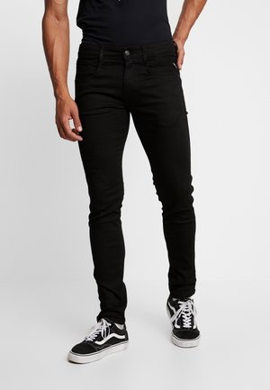 Man wearing slim-fit black jeans and black-and-white sneakers standing against a plain light background.