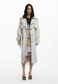 Light beige trench coat with large collar, front buttons, and belted waist. Worn over a multicolored, patterned dress and black strappy heels.