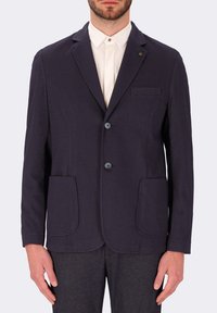 Blazer blu Navy con texture, due tasche frontali, chiusura a due bottoni e reverse. Indossato sopra una camicia bianca con colletto.