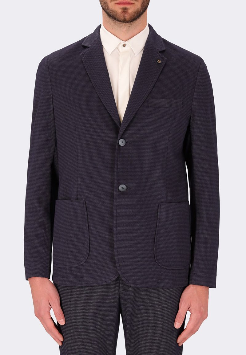 Blazer blu Navy con texture, due tasche frontali, chiusura a due bottoni e reverse. Indossato sopra una camicia bianca con colletto.