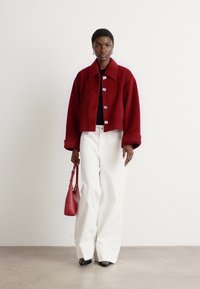 Red cropped jacket with a collar and button closure, paired with wide white trousers. Holding a small red handbag and wearing black shoes.