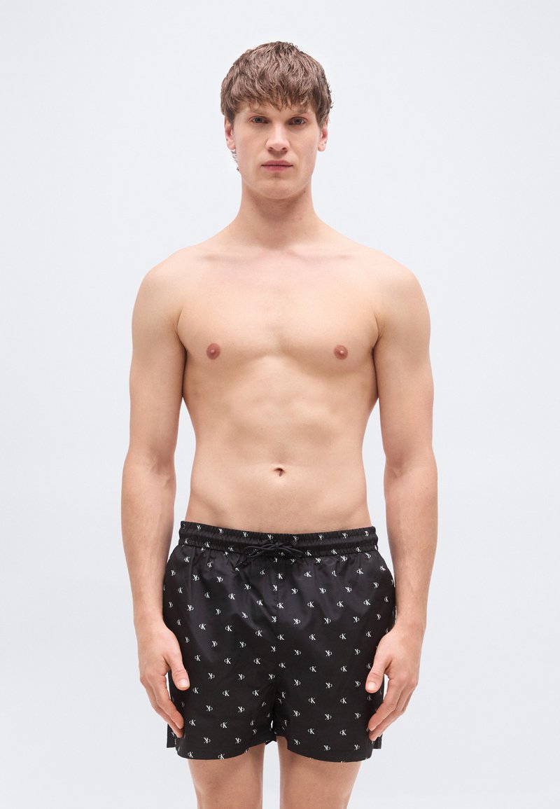Ung mann som står vendt mot betrakteren, skjorteløs, iført svarte badeshorts med hvitt "CK" monogrammønster og snor i livet.