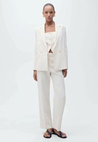 Crème-kleurig driedelig pak met een fitted top, brede broekspijpen en een bijpassende blazer met gouden knoopaccenten en een gladde textuur.