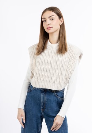 Helle beige Strickjacke mit Rippstruktur, übergroßem Design und hohem Kragen, getragen über einem weißen Langarmhemd und blauen Jeans.