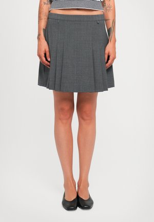Person som har på seg en grå plisserte skjørt, svarte sko, stripete crop top, med synlige tatoveringer på armene mot en ensfarget hvit bakgrunn.