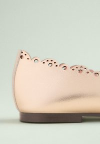 Chaussure en cuir rose pâle avec un bord festonné et des détails perforés, semelle plate marron, présentant une texture lisse et une forme arrondie.