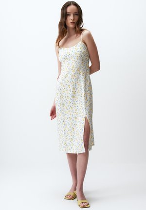 Vestito midi bianco a fiori con motivi gialli e blu, spalline sottili, spacco laterale e tessuto leggero. La modella indossa sandali verdi.