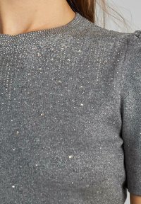 Szary, dzianinowy top o chropowatej fakturze z rozrzuconymi metalicznymi akcentami. Okrągły dekolt i krótkie rękawy, dopasowana sylwetka.