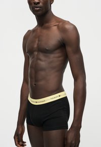 Μυώδης σκουρόχρωμος άνδρας που φοράει μαύρα boxer Tommy Hilfiger με μπεζ ζώνη, στέκεται μπροστά σε λευκό, απλό φόντο.