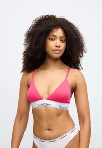 Rozā trīsstūrveida krūšturis ar plānām siksniņām, kura priekšā ir balta lenta ar "Calvin Klein" logo. Gluda tekstūra ar vienkāršu dizainu.