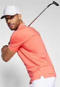 Polo shirt corallo con maniche corte, tessuto traspirante, fessure laterali, cappellino bianco e un club da golf posizionato dietro la spalla della persona.