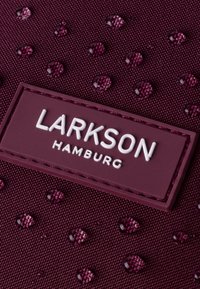 Tissu bordeaux avec des gouttes d'eau en relief et brillantes, et un patch en caoutchouc rectangulaire étiqueté "LARKSON HAMBURG" en lettres blanches.