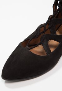 Chaussure en daim noir avec un bout pointu et un design ajouré comportant des détails de laçage. L'intérieur est en cuir beige, mettant en valeur une texture douce.