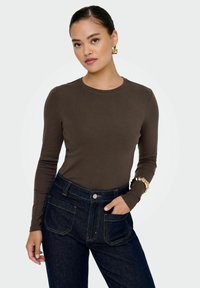 Maglione marrone a maniche lunghe con scollatura rotonda, abbinato a jeans a vita alta blu scuro. Tessuto con texture visibile; bracciale dorato in evidenza vicino al polso.