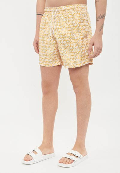 Bermuda gialle con un motivo di testo bianco ripetuto, cintura elastica e coulisse. Abbinati a sandali bianchi a scivolo.