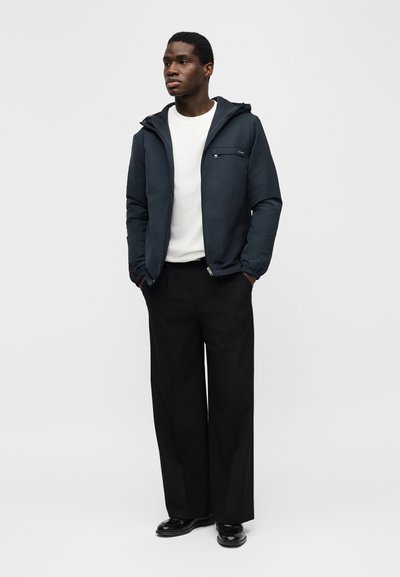 Άνδρας που φοράει ένα ναυτικό μπλε μπουφάν με κουκούλα πάνω από ένα λευκό πουκάμισο, μαύρο παντελόνι φαρδιάς γραμμής και μαύρα παπούτσια, όρθιος με τα χέρια στις τσέπες.