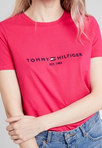 Camiseta de algodón fucsia con cuello redondo, que presenta "TOMMY HILFIGER" bordado en azul marino y "EST. 1985" debajo.