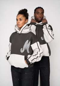 Hanorace în culoarea cărbune și alb cu buzunar frontal, brodate cu litera „M” și mâneci contrastante. Cuplul de modele stă spate în spate.