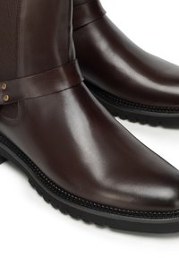 Bottes courtes en cuir marron foncé, avec une finition lisse, des panneaux élastiques latéraux et une semelle en caoutchouc texturée avec des détails sur le bord.