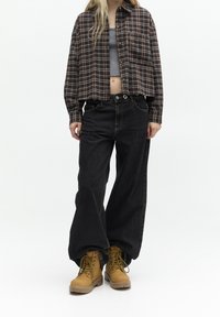 Camisa corta de cuadros marrones y negros sobre una camiseta gris, combinada con pantalones vaqueros negros holgados y botas beige con cordones. Fondo minimalista.