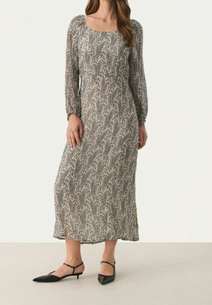 Frau trägt ein langärmeliges, knöchellanges Kleid mit einem schwarz-weißen Paisley-Muster und schwarze spitze Schuhe mit niedrigem Absatz.