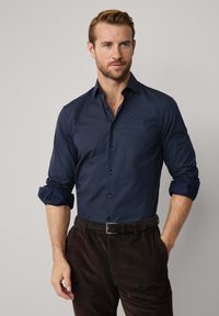 Man in een donkerblauw, gemêleerd overhemd met opgerolde mouwen en donkere bruine broek, staand met één hand in zijn zak.