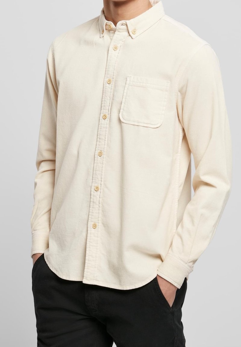 Homme portant une chemise crème à manches longues avec poche poitrine et pantalon noir, les mains dans les poches, sur un fond uni.