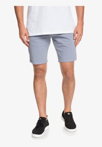 Muž v světlých šedých šortkách, bílém tričku a černých teniskách, stojící s rukama u boků.