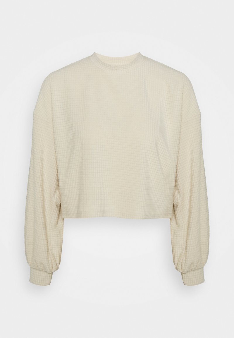 Krämfärgad kort topp med en texturerad, våffelmönster. Har långa, voluminösa ärmar och en rund halsringning.