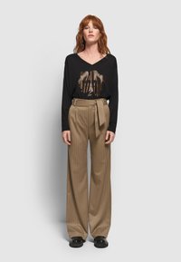 Roodharige vrouw in een zwarte langmouw T-shirt met grafische motieven, hoge beige gestreepte wijde broek, en zwarte schoenen, staand.