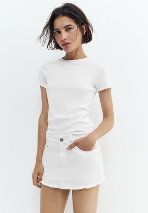 Femme aux cheveux foncés portant un t-shirt blanc uni et une mini-jupe en denim blanche, une main dans la poche, debout devant un fond blanc.
