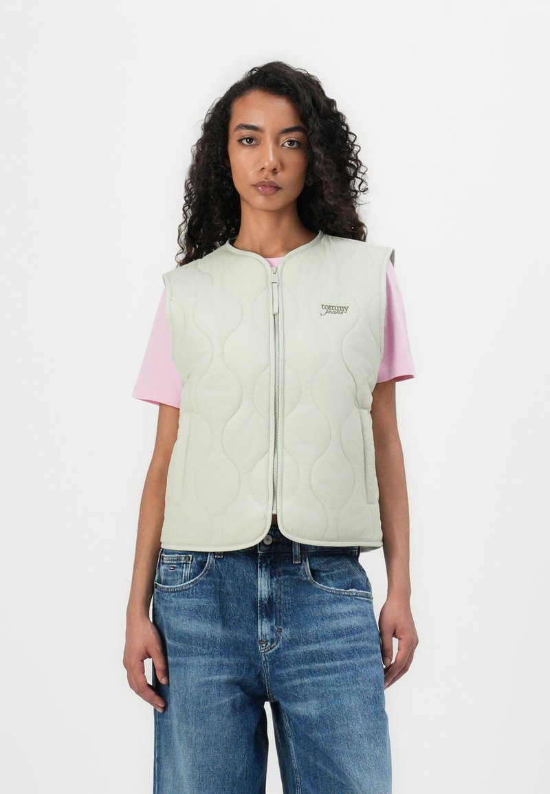 Gilet matelassé vert pâle avec un col rond et une fermeture éclair à l'avant. Présente un logo brodé et des manches courtes sur une chemise rose et un jean en denim.