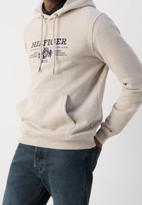 Sudadera de algodón beige con un bolsillo frontal, capucha con cordón y detalles del logotipo en azul marino. Combinada con unos vaqueros de denim azul oscuro.