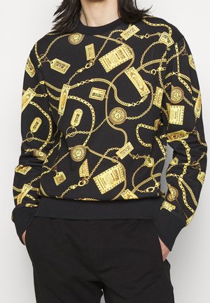 Sweat-shirt noir avec impression de chaîne et d'étiquette dorées, porté par une personne les mains dans les poches d'un pantalon noir, ne montrant que le haut du torse.