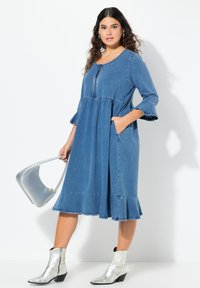 Denim-mekko, jossa on pyöreä pääntie, kolmivartinen kellohelma ja levenevä helma. Edessä on vetoketju ja sivutaskut. Yhdistetty hopeisiin saappaisiin.